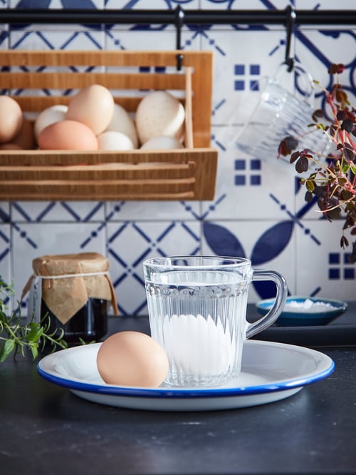Close-up of a DRÖMBILD mug in clear glass with an egg inside, sitting on a blue and white plate on a kitchen countertop.