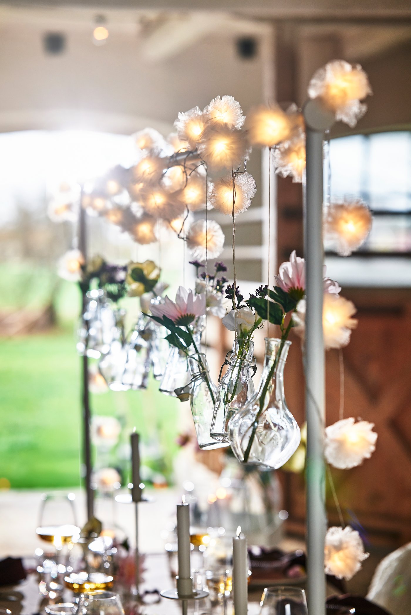 Festlich gedeckter Tisch mit hängenden Lichterketten und Glasvasen mit Blumen, Kerzen und Gläsern, im Hintergrund ein Garten.