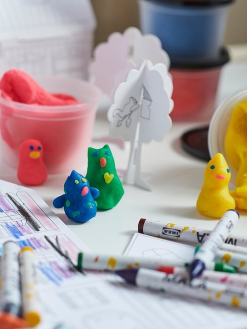 A tabletop filled with MÅLA wax crayons, figures of MÅLA mixed-colour modelling dough, and MÅLA cardboard town templates.