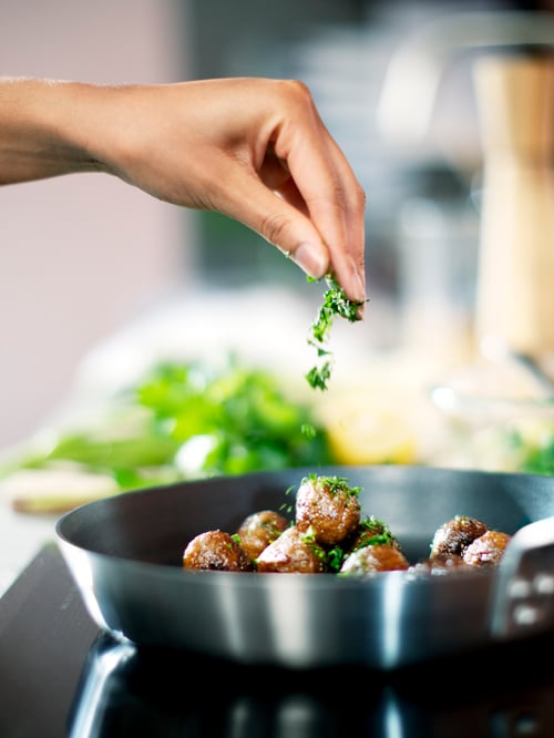 Bucătărie cu plită, accesorii de gătit, ingrediente, chiftele într-o tigaie, tocător și o mână presărând ierburi verzi.