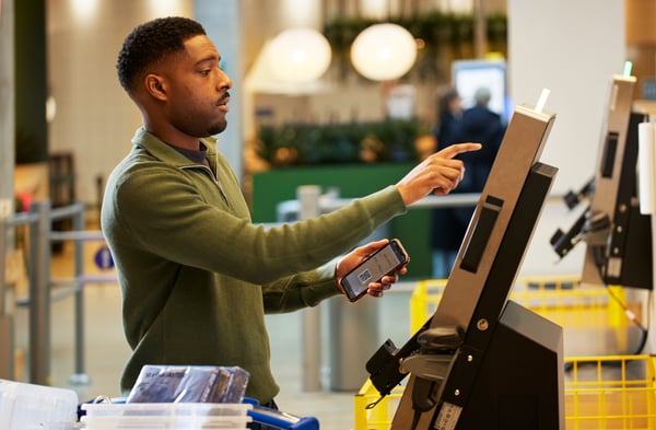 Person använder en självbetjäningsskärm i butik och håller en mobiltelefon i handen.