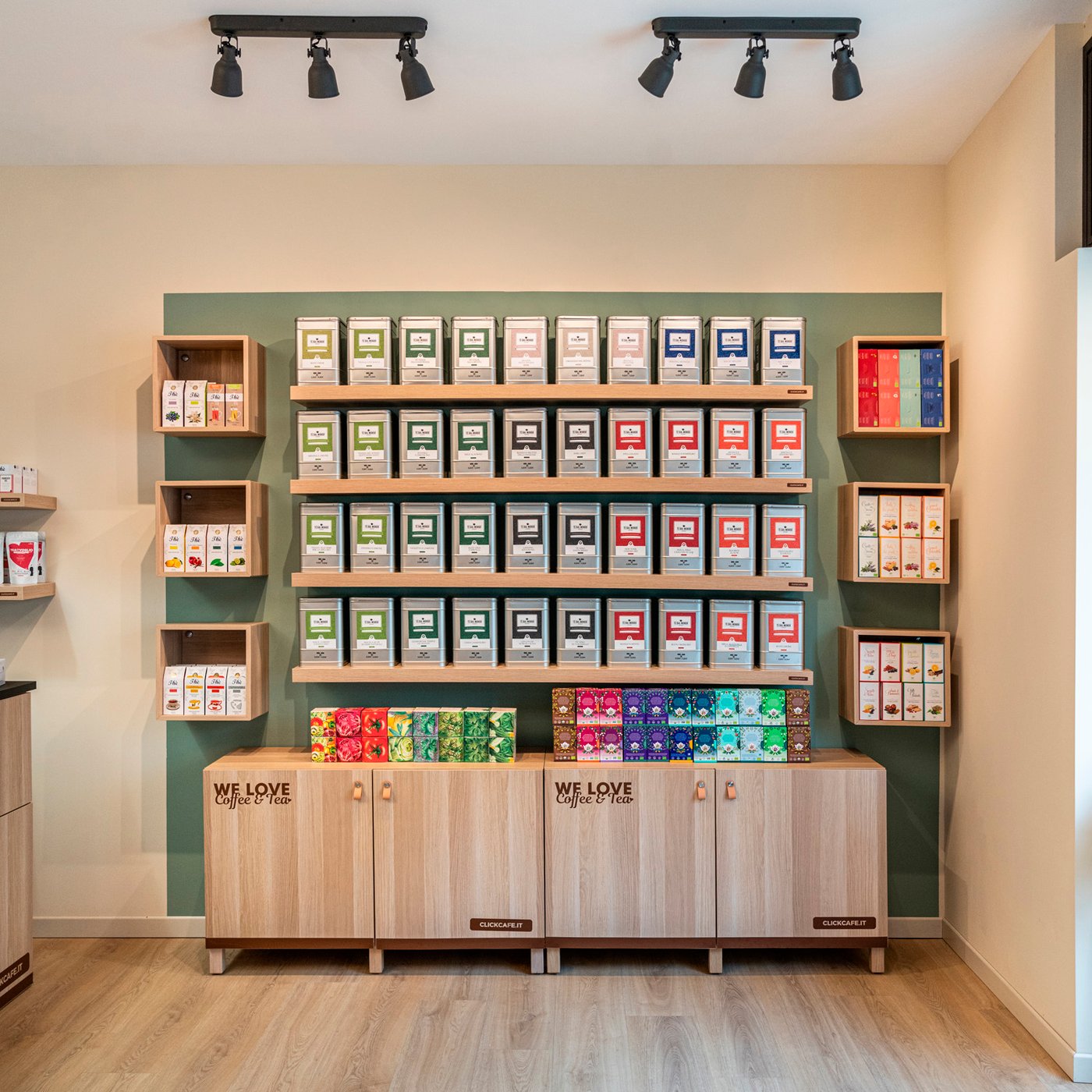 Una parete attrezzata con scaffali di un negozio di tè e caffè. I mobili sono tutti in impiallacciatura di rovere mordente bianco. La parete è crema con un quadrato verde oliva e all'interno ci sono tutti i mobili a parete.