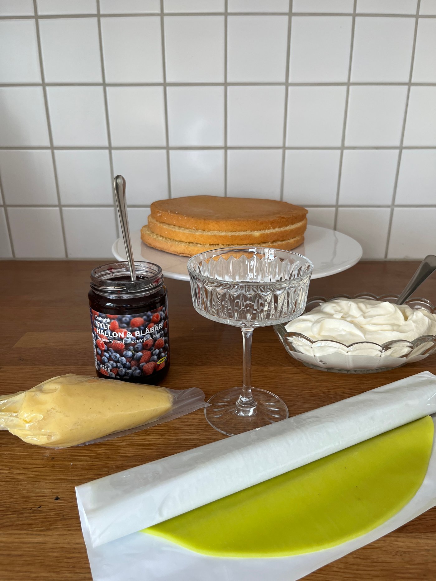 Ekologisk SYLT HALLON & BLÅBÄR hallon- och blåbärssylt, ett SÄLLSKAPLIG glas, marsipantäcke, vispad grädde i en skål och tårtbottnar står på en köksbänk.