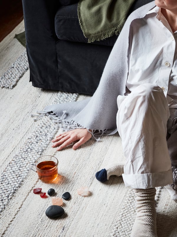 En rødhåret kvinne med et sjal over skuldrene sitter på et gulvteppe og lener seg mot en sofa. Foran seg har hun et glass te og noen steiner.