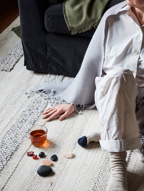 Partial shot of a person sitting on a rectangular low pile rug. On the rug is a mug and multiple coloured rocks.