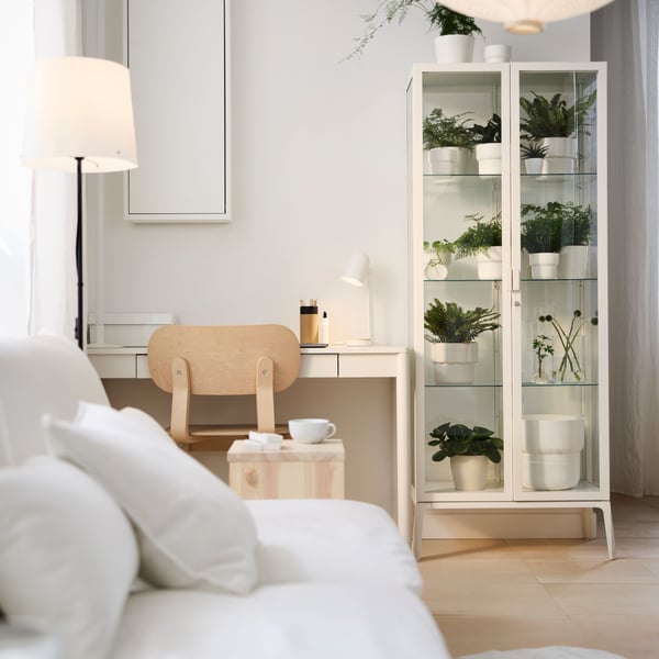 A white TONSTAD desk and a chair create a workspace in a multifunctional room with plants in a MILSBO glass-door cabinet.