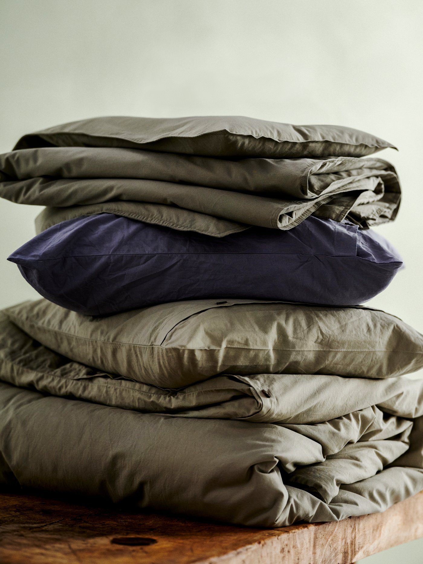 Ropa de cama doblada una encima de otra sobre una tabla de madera, con una funda de plumón y una funda de almohada KRÅKRISMOTT en verde claro en el medio. 