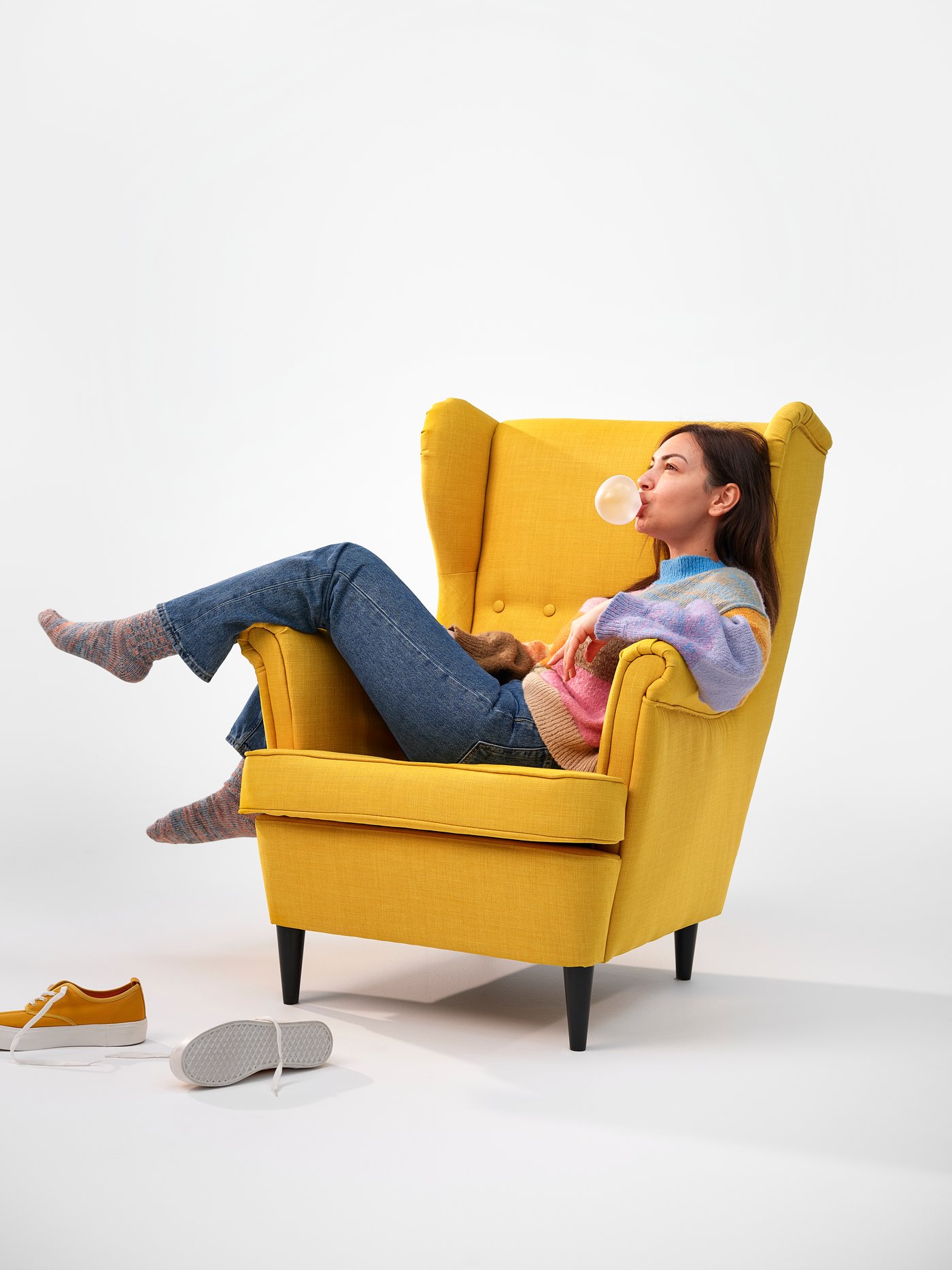 A person lounging across the arms of a comfy-looking yellow wing chair in a white space.