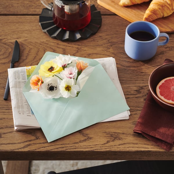 The heads of a number of GULDSPETT artificial flowers poke out of an envelope near a TOBISFISK mug on top of a table.