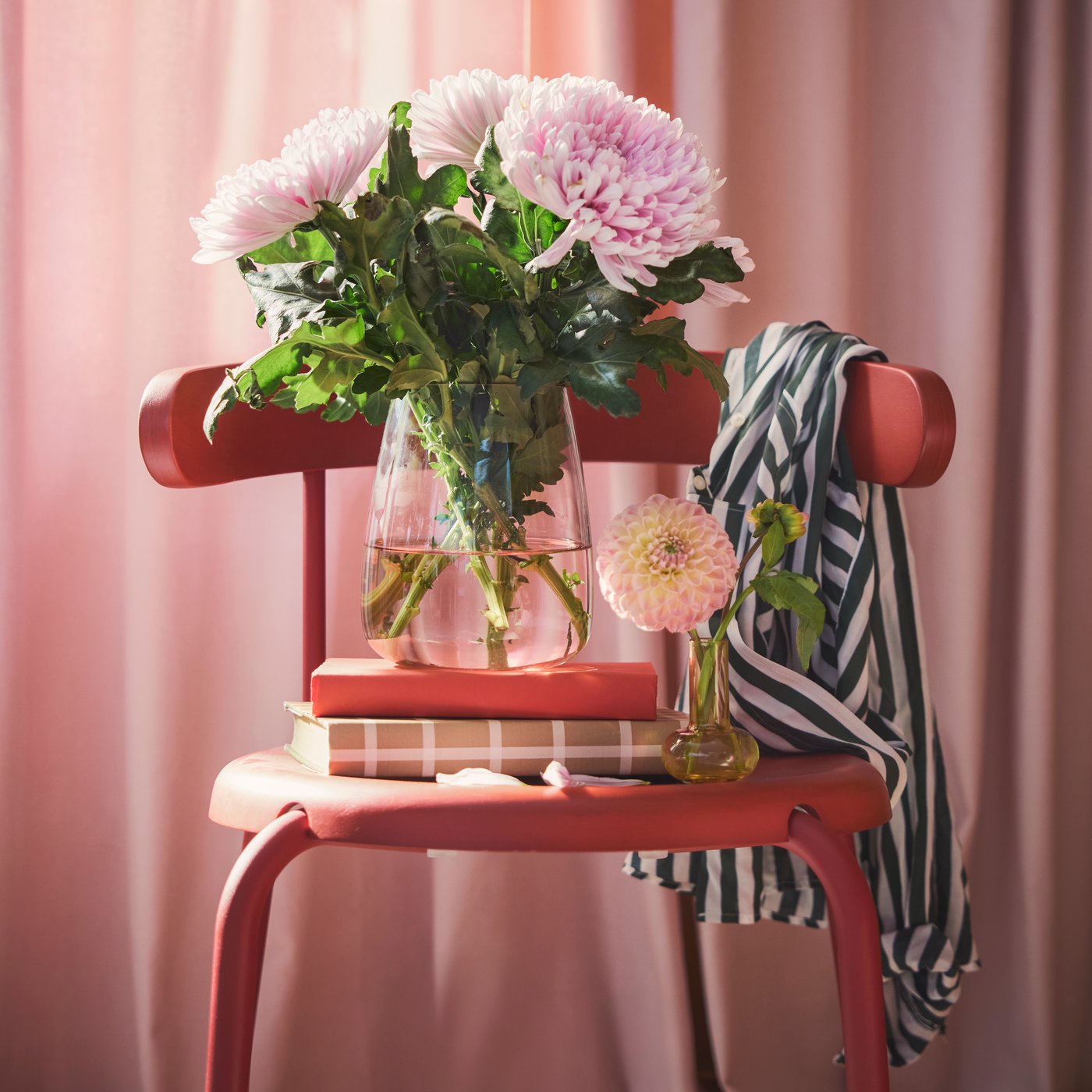 A living room features a light pink BERÄKNA vase made of glass filled with large pink flowers on a red chair beside books..