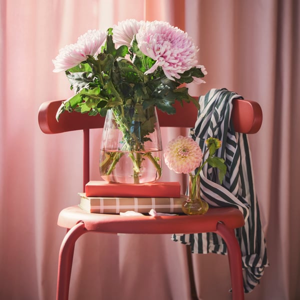 A living room features a light pink BERÄKNA vase made of glass filled with large pink flowers on a red chair beside books.