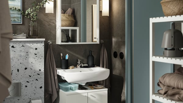 Une petite salle de bain aux murs brun foncé et aux meubles blancs. Un élément NYSJÖN à deux portes sert de rangement sous la vasque.