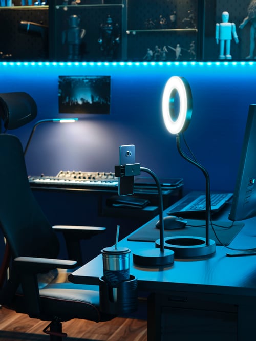 Eine LÅNESPELARE Ringleuchte mit Mobiltelefonhalterung auf einem Schreibtisch in einem blau beleuchteten Raum. Im Vordergrund steht ein MATCHSPEL Gamingstuhl.