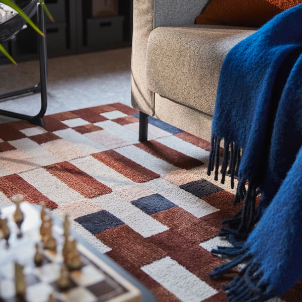 Un salón con una alfombra de pelo corto MARKBLÅMME multicolor con un estampado de cuadros debajo de un sofá y junto a un juego de ajedrez.