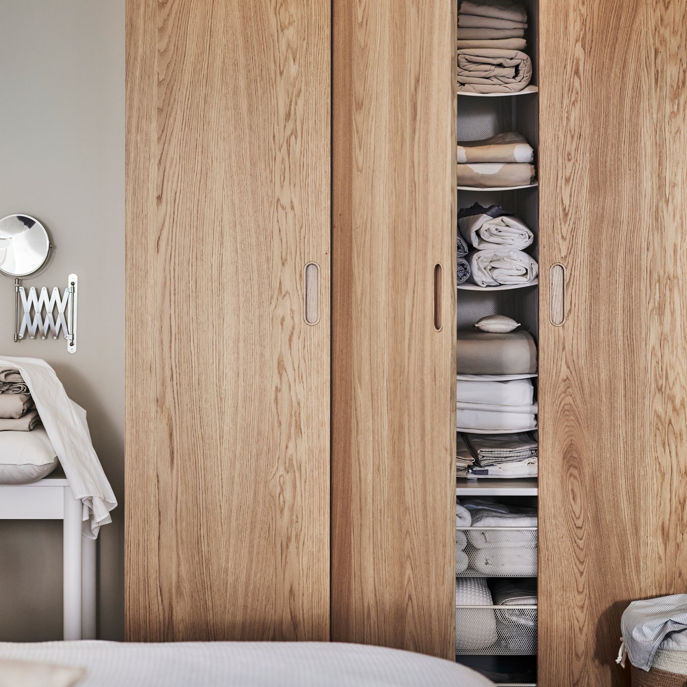 Ett träskåp med skjutdörrar där en dörr är öppen och visar organiserade hyllor med vikta handdukar, lakan och textilier. Bredvid skåpet står ett litet bord med vikta tyger.
