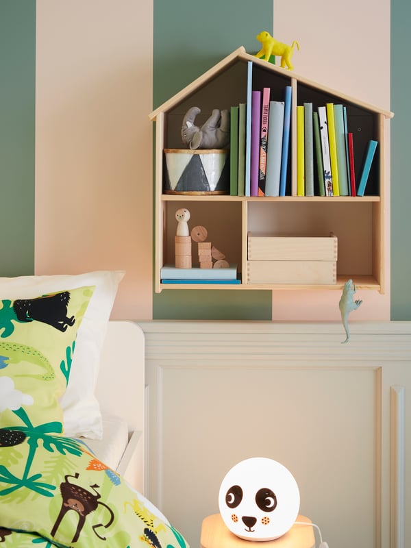 A FLISAT doll’s house/wall shelf with books and toys is mounted on the wall above an ÄNGARNA LED table lamp near a SLÄKT bed.