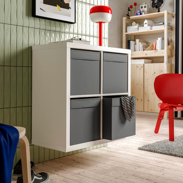 Deel van een wand met een wandgemonteerde witte KALLAX stellingkast met een donkergrijze DRÖNA doos in elk van de vier vakken.