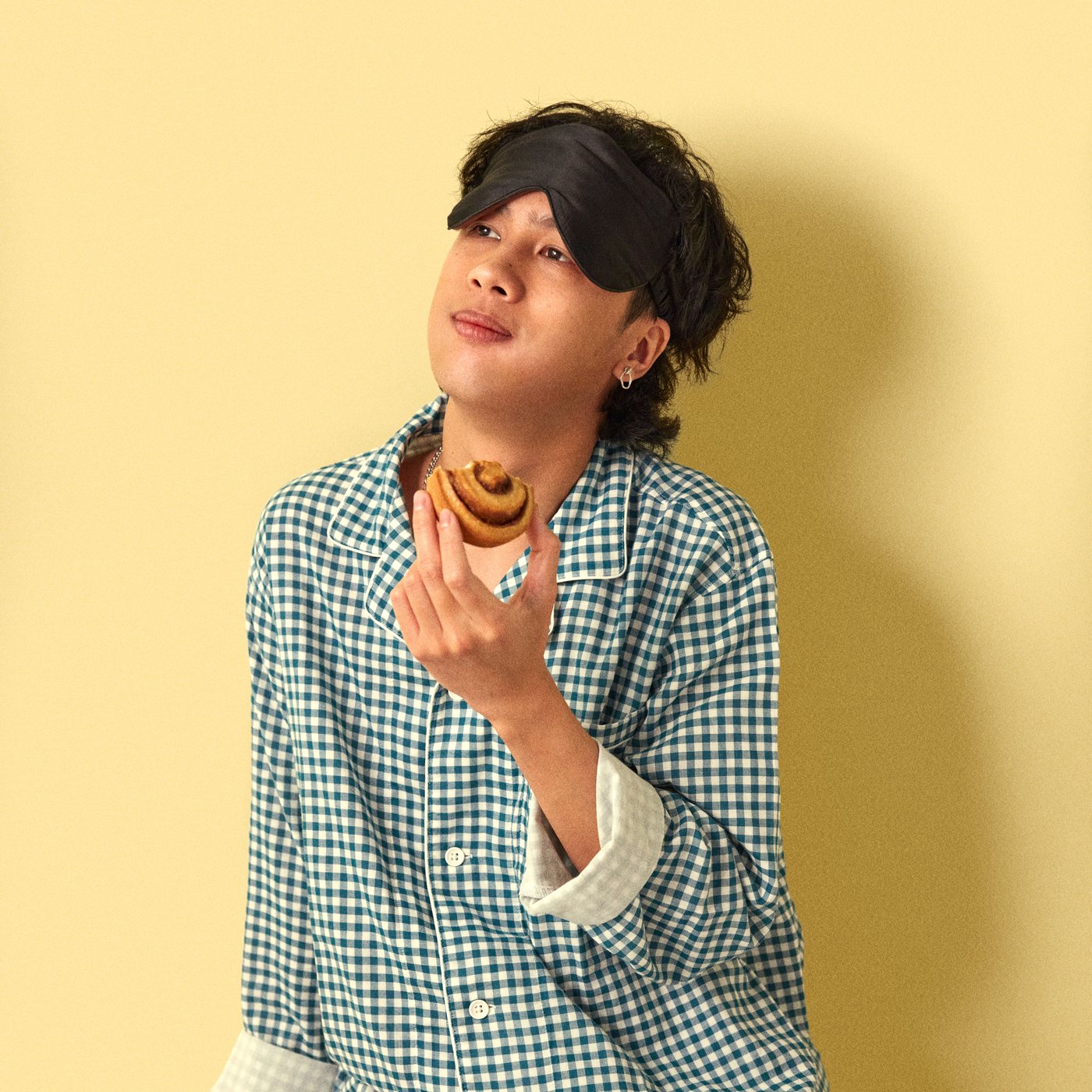 A young man wearing a green pyjama is eating a cinnamon bun against a yellow wall.