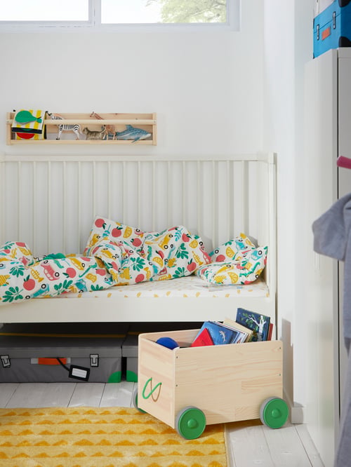 Child's bedroom with white wooden cot, colourful bed linens, yellow rug, wooden shelves and wooden toy storage box.