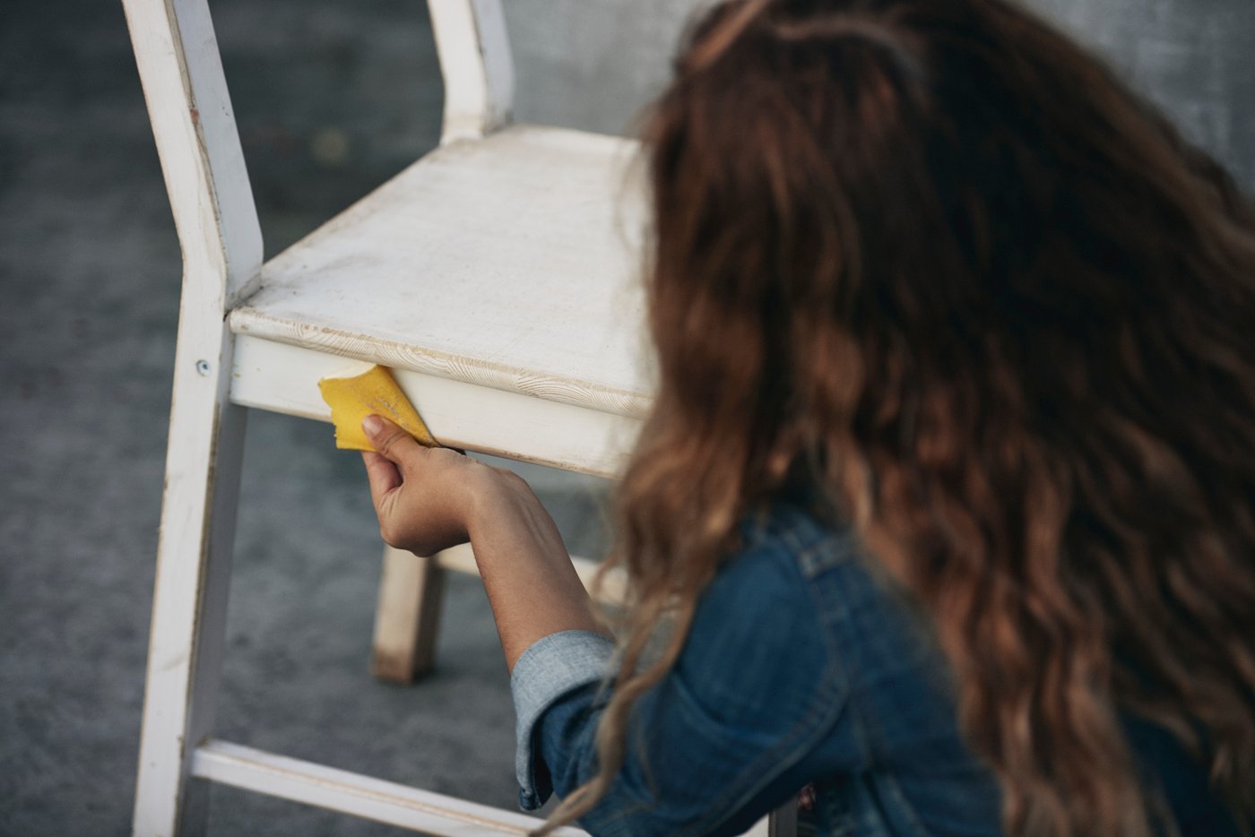 Людина з довгим каштановим волоссям наждачним папером шліфує пофарбований білим дерев'яний стілець, що стоїть на сірій підлозі.