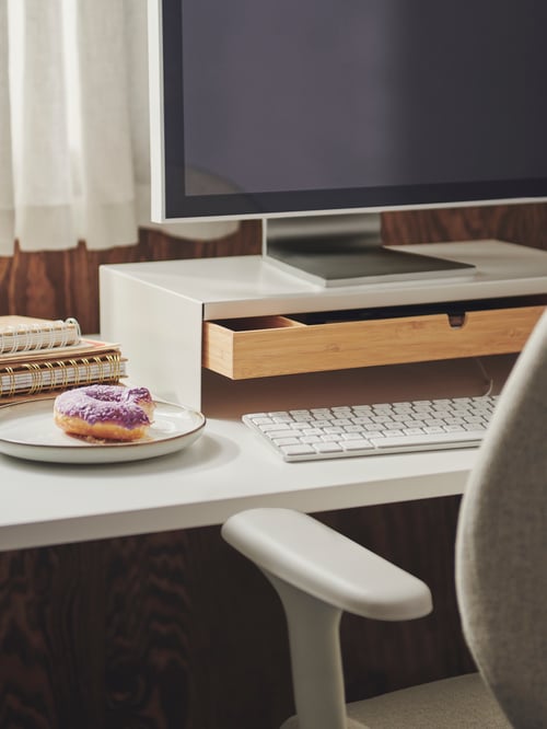 Close-up of workspace with desk, office chair, and PC monitor on top of rectangular white steel monitor stand with drawer.