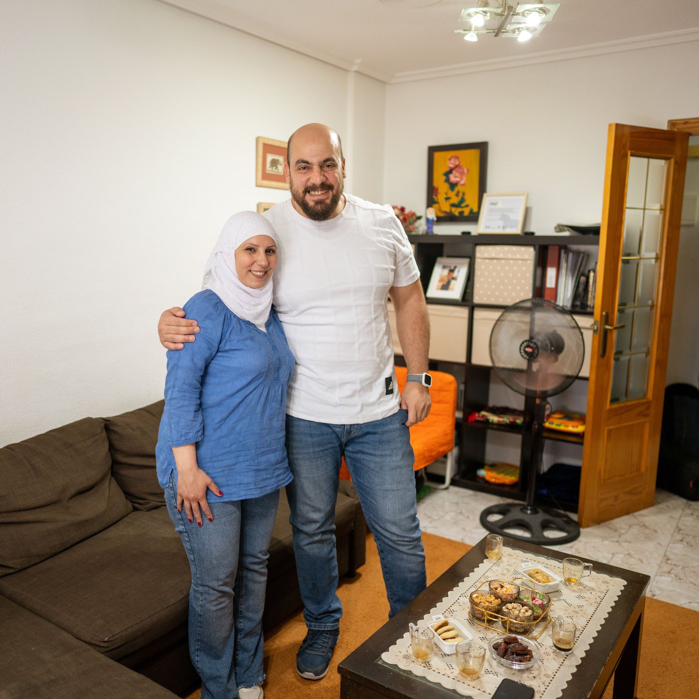 A couple stands in their living room, holding each other while smiling at the camera before having a coffe break.