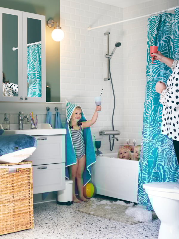 Bathroom with three girls around a bath. One girl, draped in a RÖRANDE hooded towel, makes a play toast with her mother.