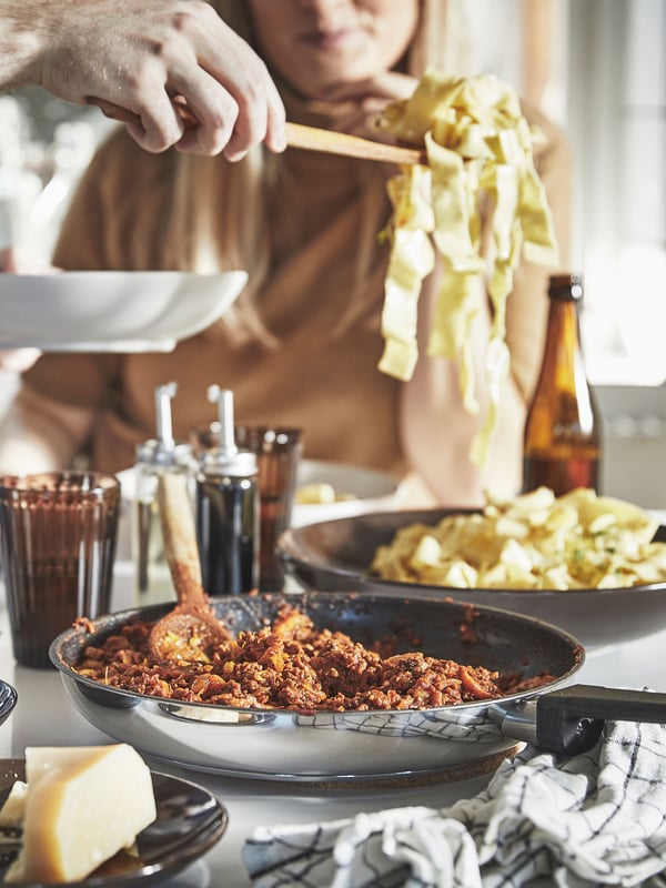 A wooden spoon is sitting in a MIDDAGSMAT frying pan filled with pasta sauce. Next to it, someone is lifting a spoonful of pasta using one hand while holding a GODMIDDAG bowl in the other hand. In the foreground, a block of cheese is on a FÄRGKLAR dish.