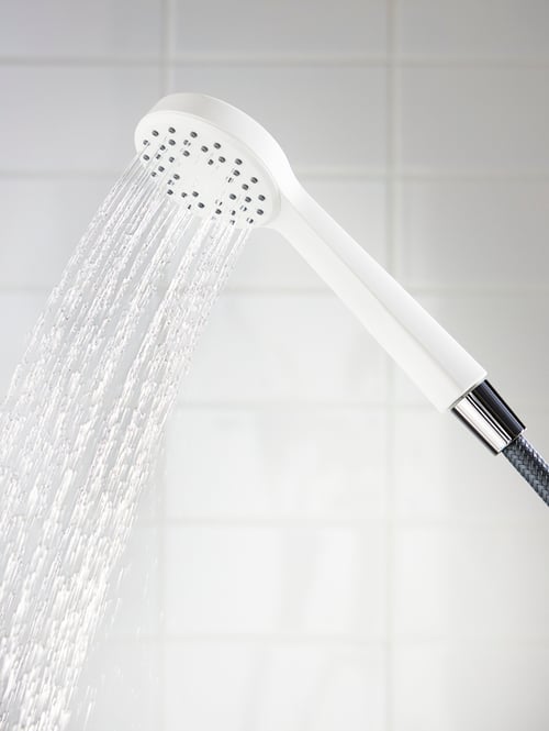 A white LILLREVET handshower spraying water, with a white-tiled wall in the background.