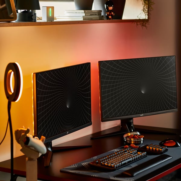 Gaming prostor z dvema zaslonoma na HUVUDSPELARE gaming mizi. Steno osvetljuje rdečerumena svetloba, ki prihaja izza zaslonov.