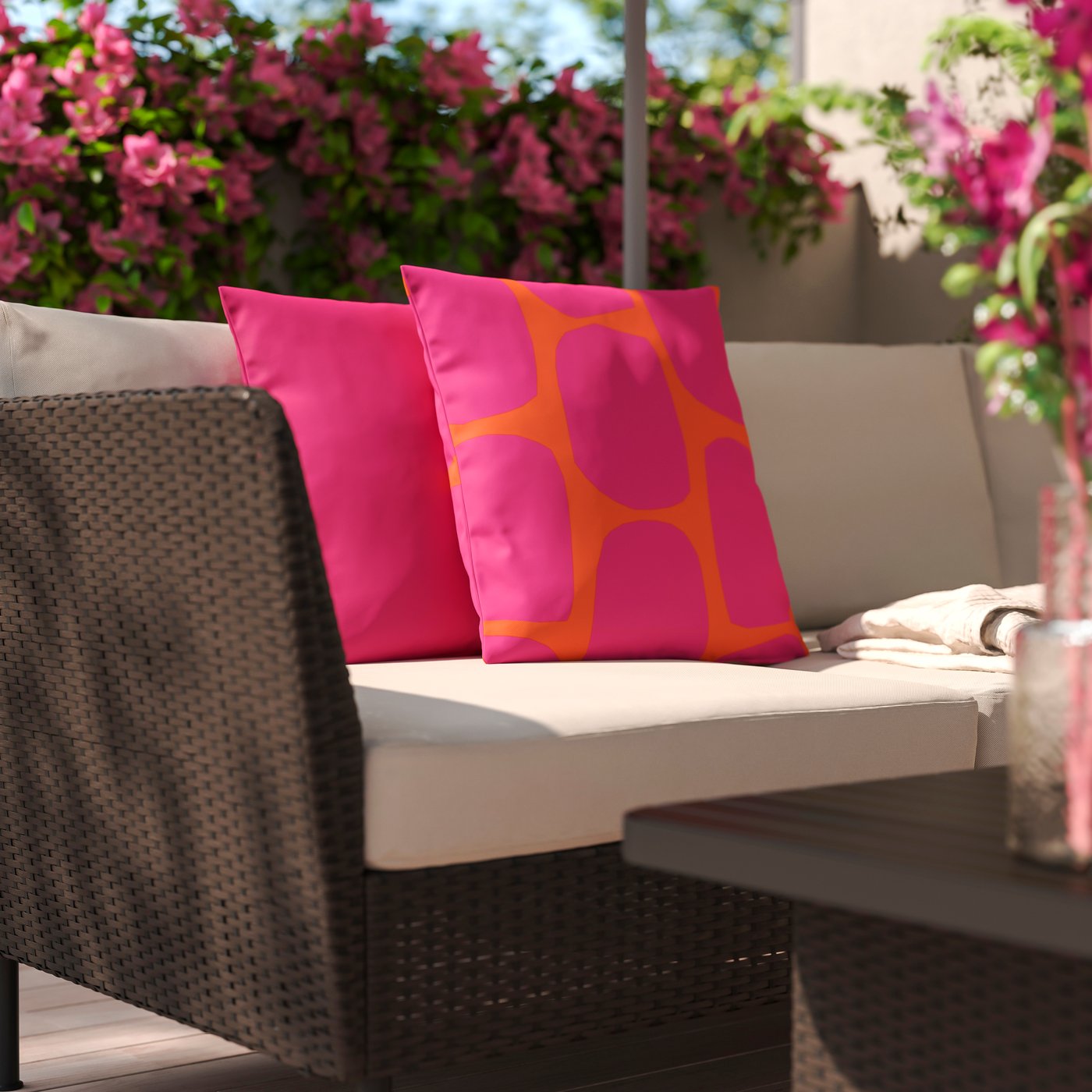 Un coin salon d'extérieur plein de vie signé IKEA, composé d'un canapé confortable aux coussins clairs, agrémenté de deux coussins décoratifs aux motifs roses et orange éclatants, le tout sur fond d'une profusion de fleurs roses.