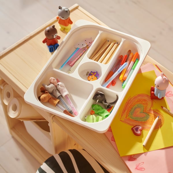 A white TROFAST tray with compartments filled with MÅLA pens and pencils on top of a TROFAST tabletop with a drawing nearby.
