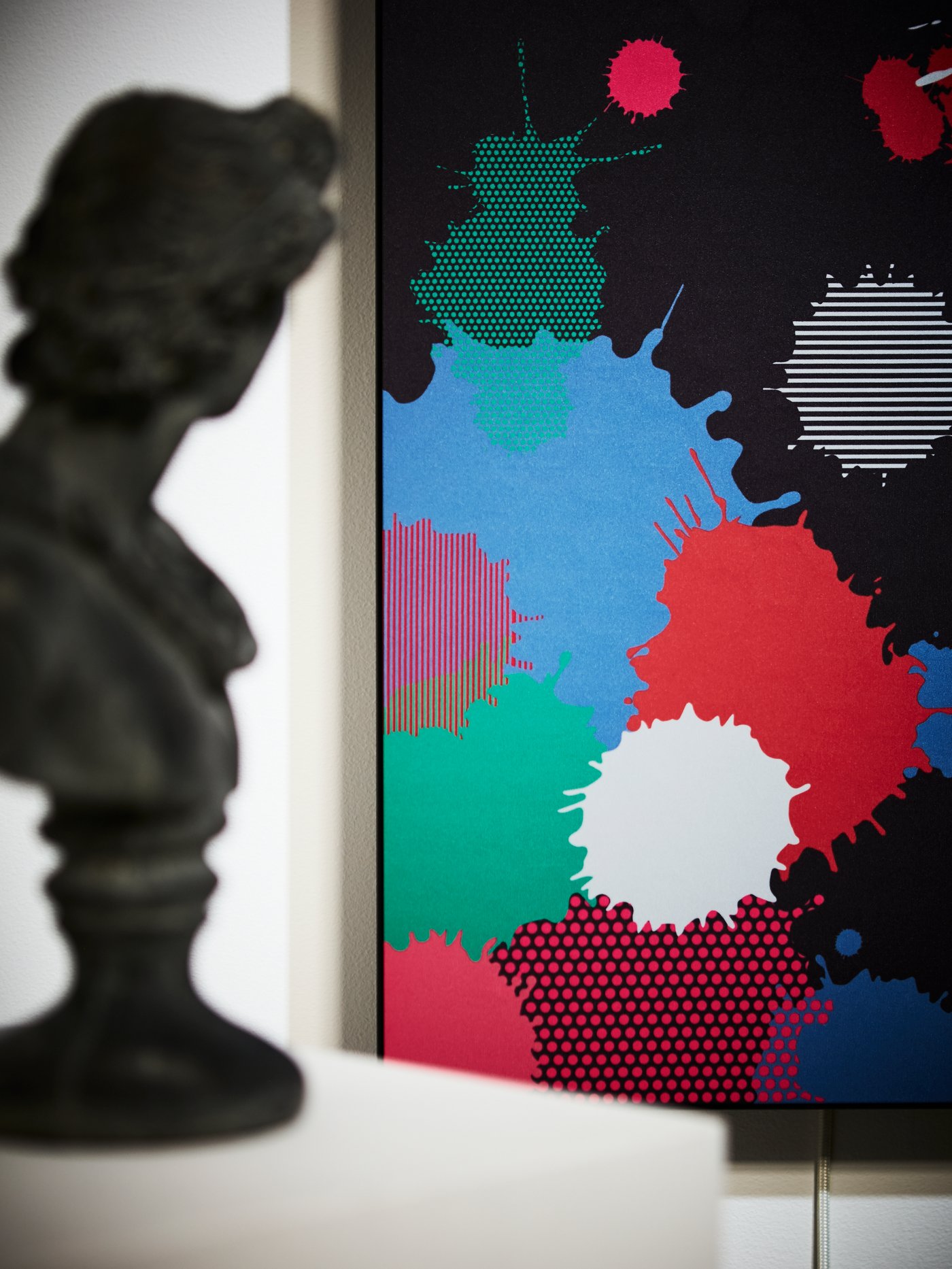 Close-up of SYMFONISK panel for picture frame speaker with paint splatter design; a bust is blurred in the foreground.