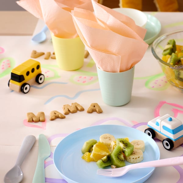 A table with pastel colored KALAS plates and cutlery, plastic cups with napkins and “mama” spelled out with letter biscuits.