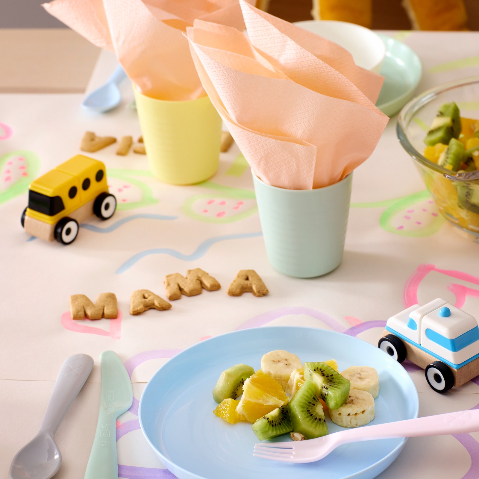 A table with pastel coloured KALAS plates and cutlery, plastic cups with napkins and “mama” spelled out with letter biscuits.