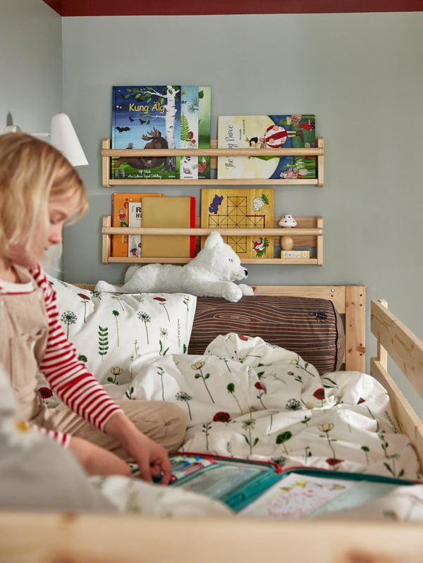Es ist eine Nahaufnahme eines Kindes zu sehen, das im oberen Bett des MYDAL Etagenbettgestells sitzt. Hinter ihr hängen FLISAT Wandaufbewahrungen mit Büchern.