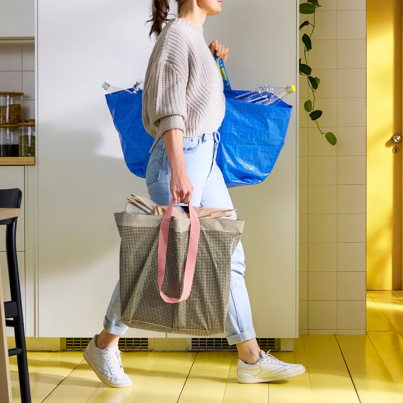 Person walking through a bright kitchen carrying two large bags, one blue and one beige, filled with various household items.