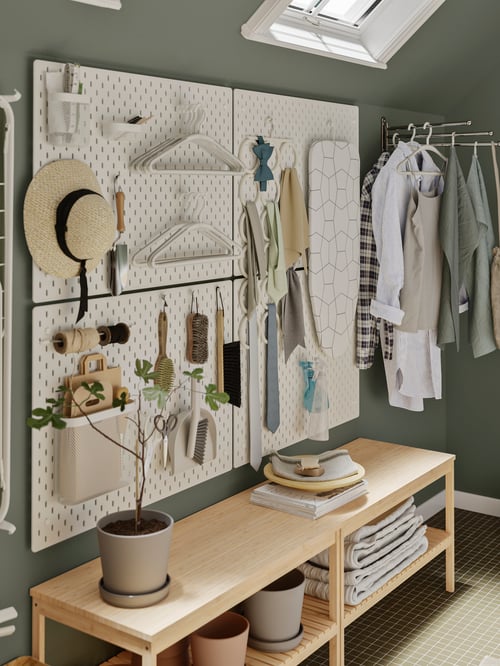 Pegboards SKÅDIS cu coșuri de depozitare, cârlige și umerașe sunt montate deasupra unei bănci RÅGRUND și lângă haine pe umerașe.