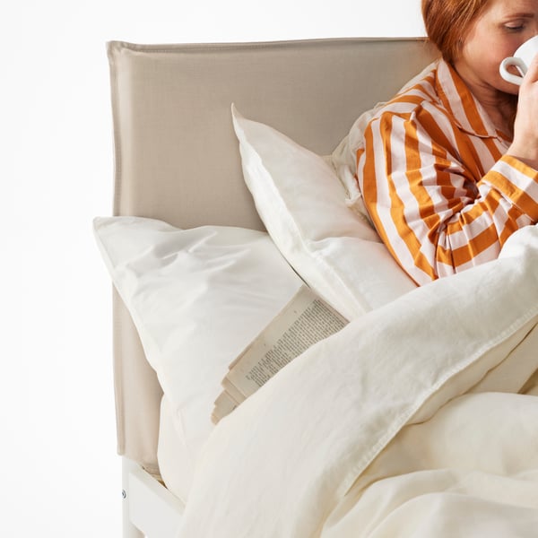 Close-up of the KLEPPSTAD bed frame’s beige fabric headboard. A person resting against the headboard is sipping from a mug.