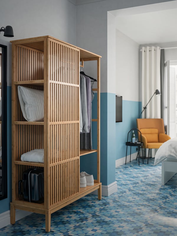A bamboo NORDKISA wardrobe with some clothes, a suitcase and an extra pillow inside. In a blue and white bedroom.
