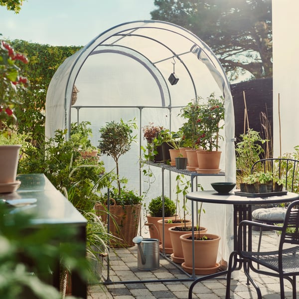 A garden with an ASPAREN greenhouse in transparent white, surrounded by potted plants and outdoor seating.