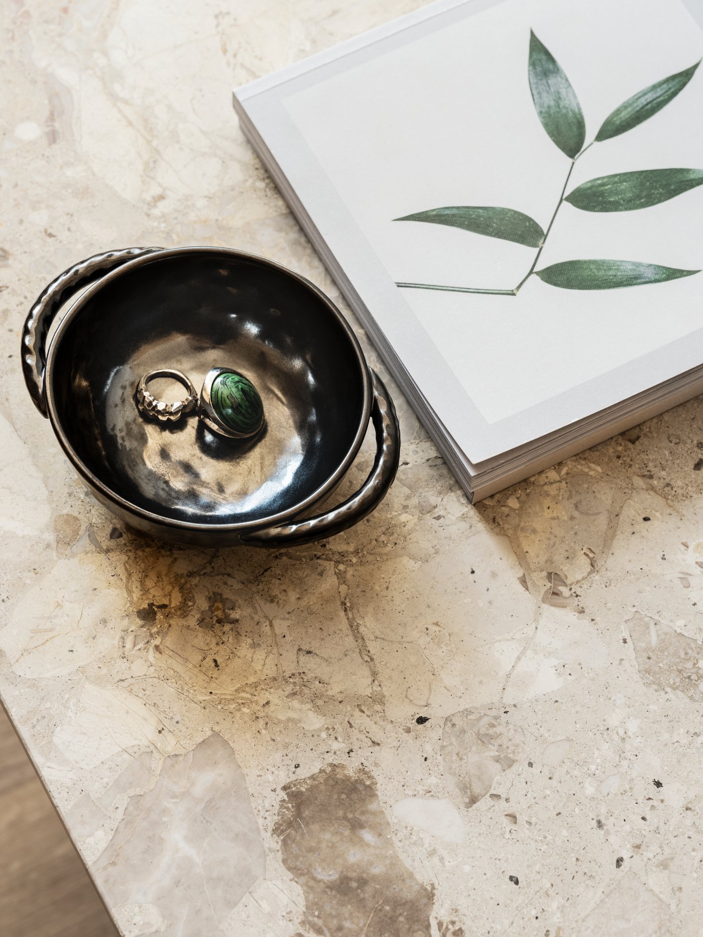 Two rings placed in a black LOKALT ceramic bowl, sitting on a marble surface next to a book with a leaf on the cover.