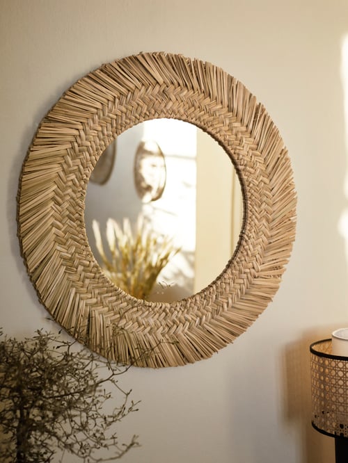 Close-up shot of a round mirror with woven fibre edges mounted on a wall, reflecting a partial section of a room.