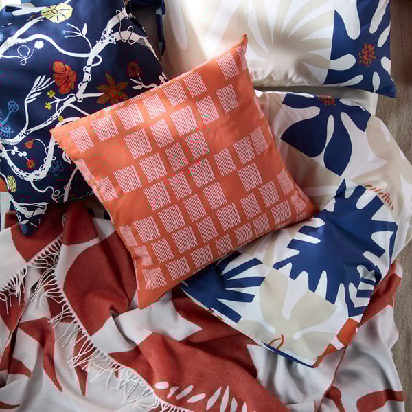 Two cushions resting on a bed with colourful bed linen and a throw. One of them has an orange PALPLJUNGMAL cushion cover.
