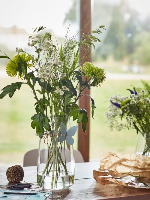 A glass vase with water and multiple flower stems is placed on a table next to paper, with another vase to the side.