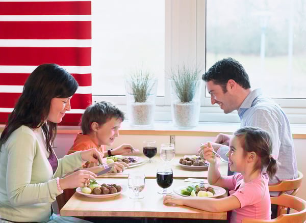 Sur cette image, une famille de quatre personnes est attablée et partage un repas dans une ambiance chaleureuse. Les parents sont assis en face l’un de l’autre, avec leurs deux jeunes enfants entre eux. Tous semblent savourer un plat à base de boulettes, probablement dans un restaurant IKEA, reconnaissable à son style épuré et à la décoration (rayures rouges et blanches, plantes en pot, grande baie vitrée). Le moment est convivial et reflète un instant de complicité familiale.