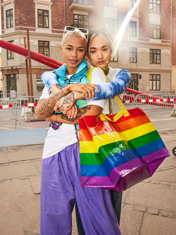 Two people sitting on the ground outside, one of them has a large STORSTOMMA bag, IKEA Rainbow Pride bag on their shoulder. 