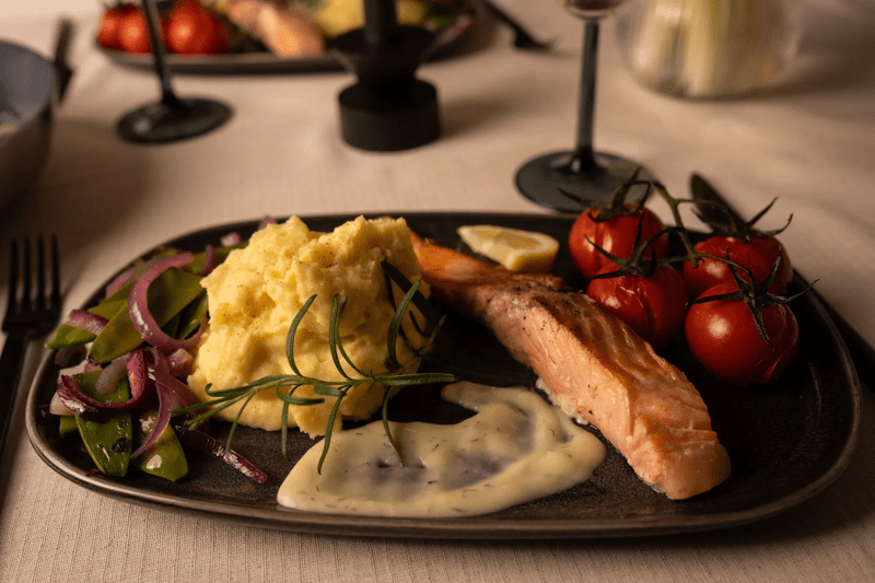 Ein Lachs mit Kartoffelpüree, Senfsauce und Gemüse auf einem dunklen Teller präsentiert.