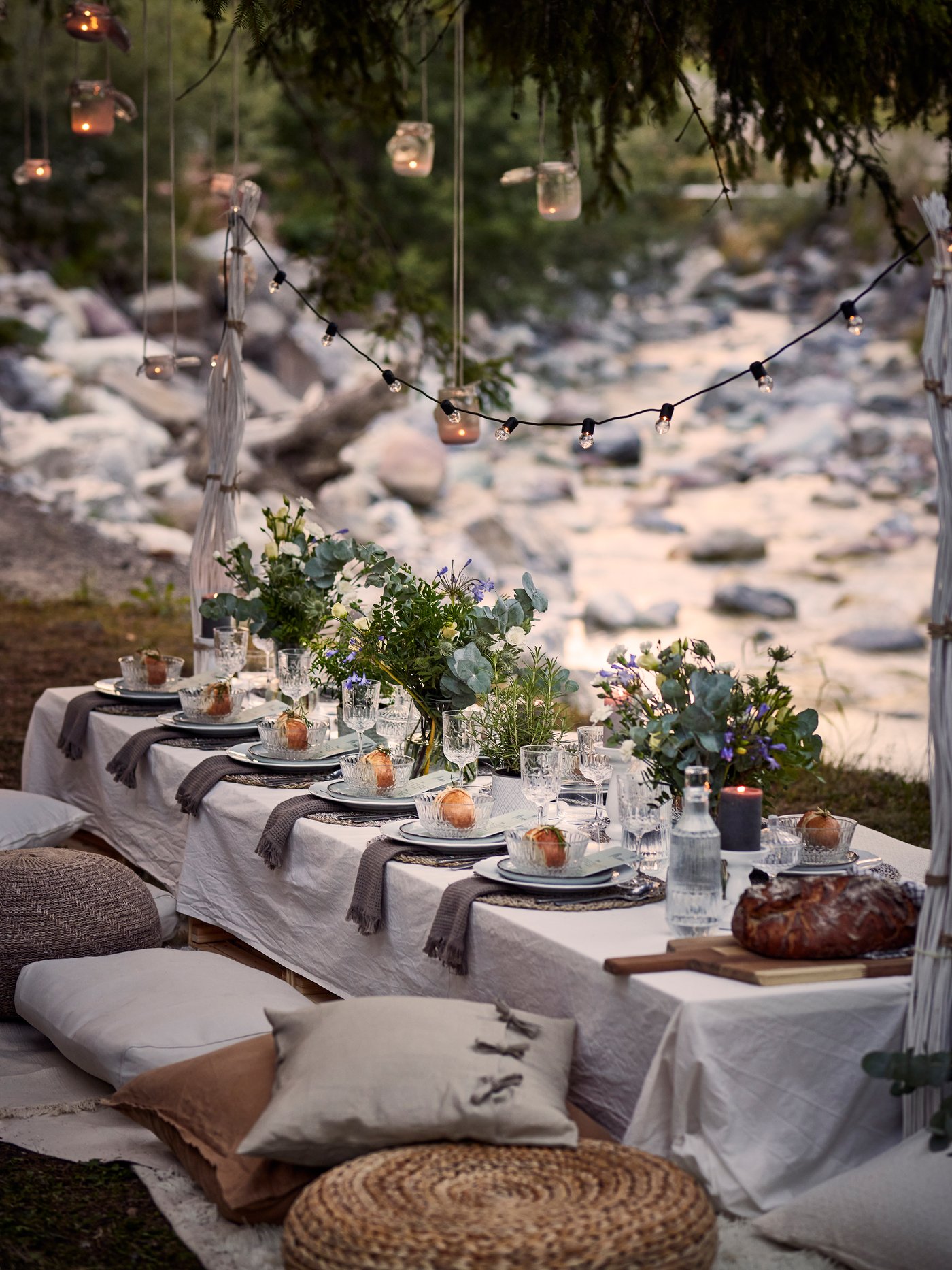 Utomhusdukning med elegant dekorerat bord täckt av vita dukar, grå servetter och blomsterarrangemang. Varje plats har tallrikar, glas och små dekorativa föremål. Området är smyckat med ljusslingor och hängande burkar med ljus. Sittplatserna består av kuddar och en vävd matta på marken.