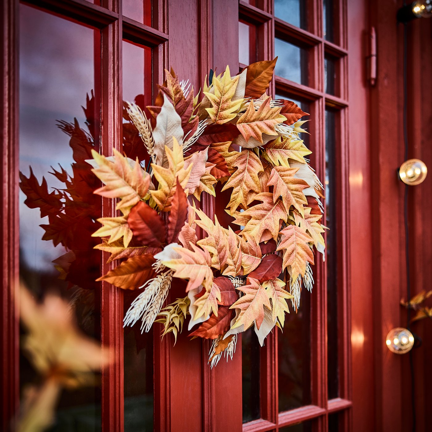 Una corona artificial HÖSTAGILLE compuesta de flores otoñales decorativas que cuelga de una puerta de entrada roja.